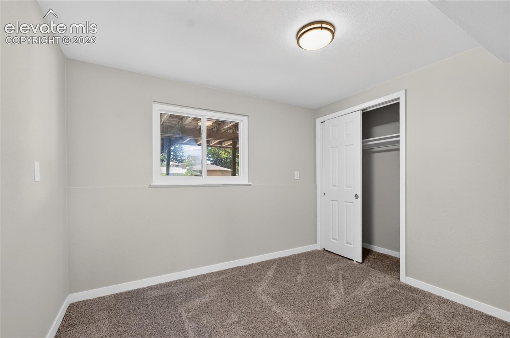 Carpeted room featuring a white-framed window, bi-fold closet doors, a ceiling-mounted light fixture, and light gray painted walls