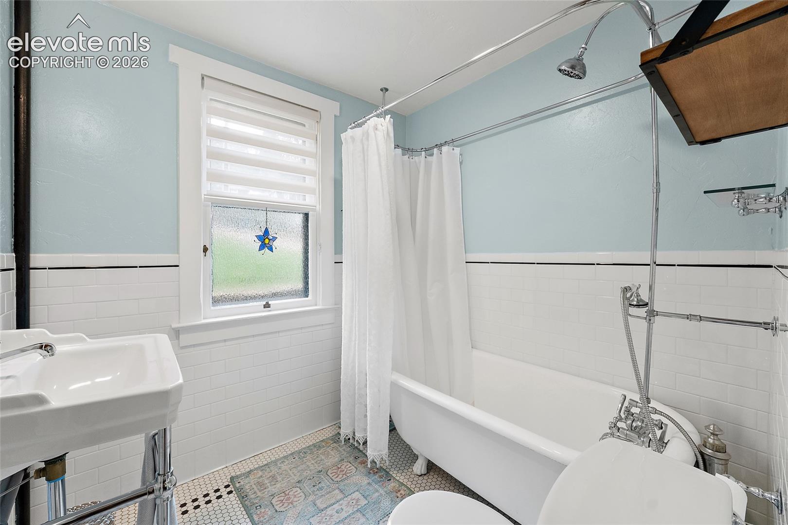 Large clawfoot tub featuring a chrome shower riser and hardware, pedestal sink, classic white subway tile wainscoting, and hexagonal mosaic tile flooring