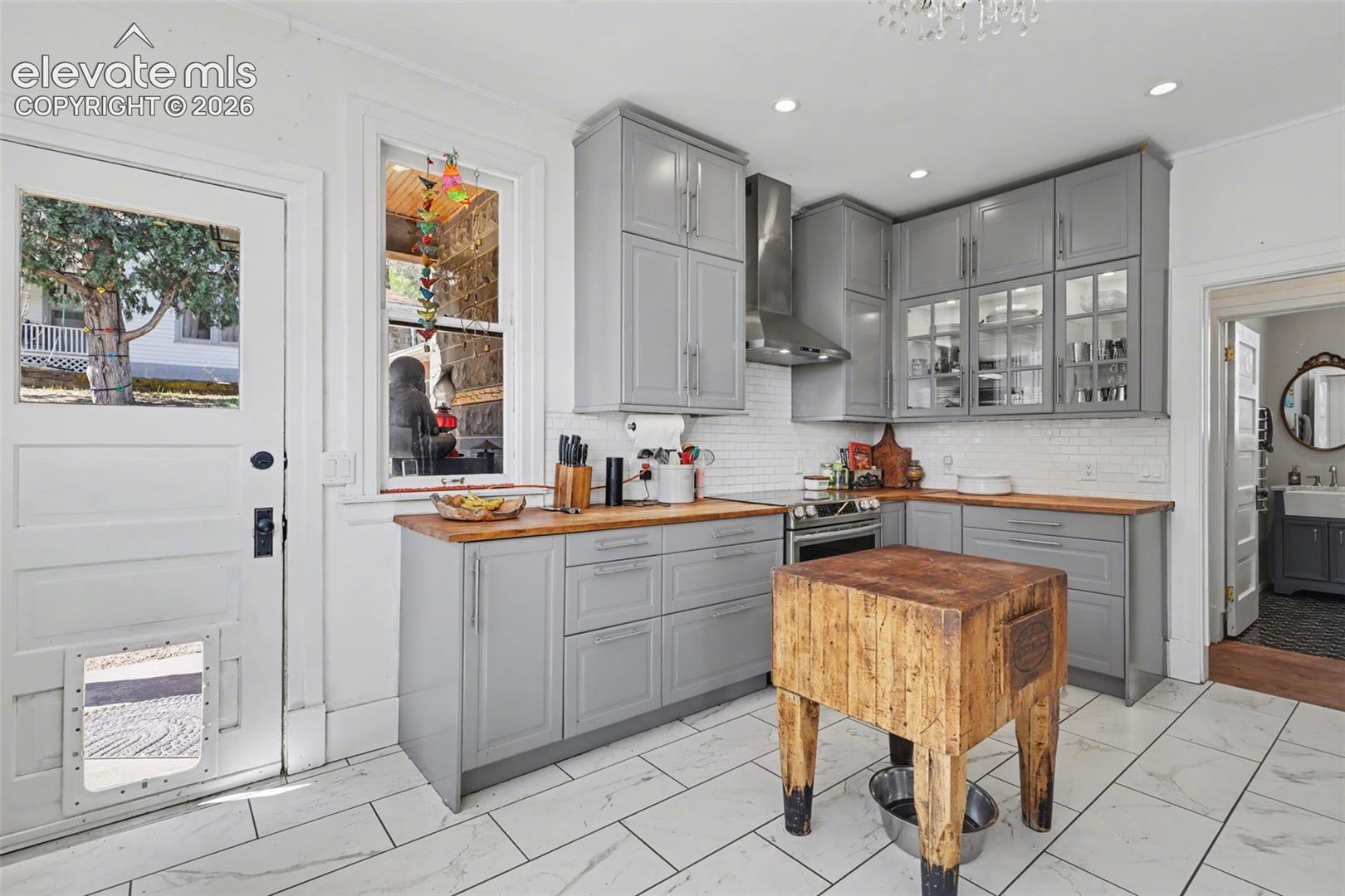 Main house kitchen featuring gray cabinets, light tile, glass insert cabinets, wood counters, and recessed lighting