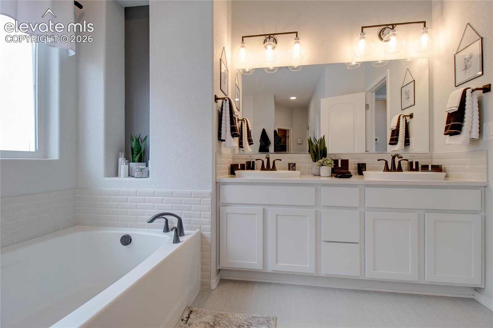 Bathroom featuring a white double-vanity cabinet with a solid surface countertop, dual vessel sinks, subway tile backsplash, and a large-format mirror with multi-light wall fixtures