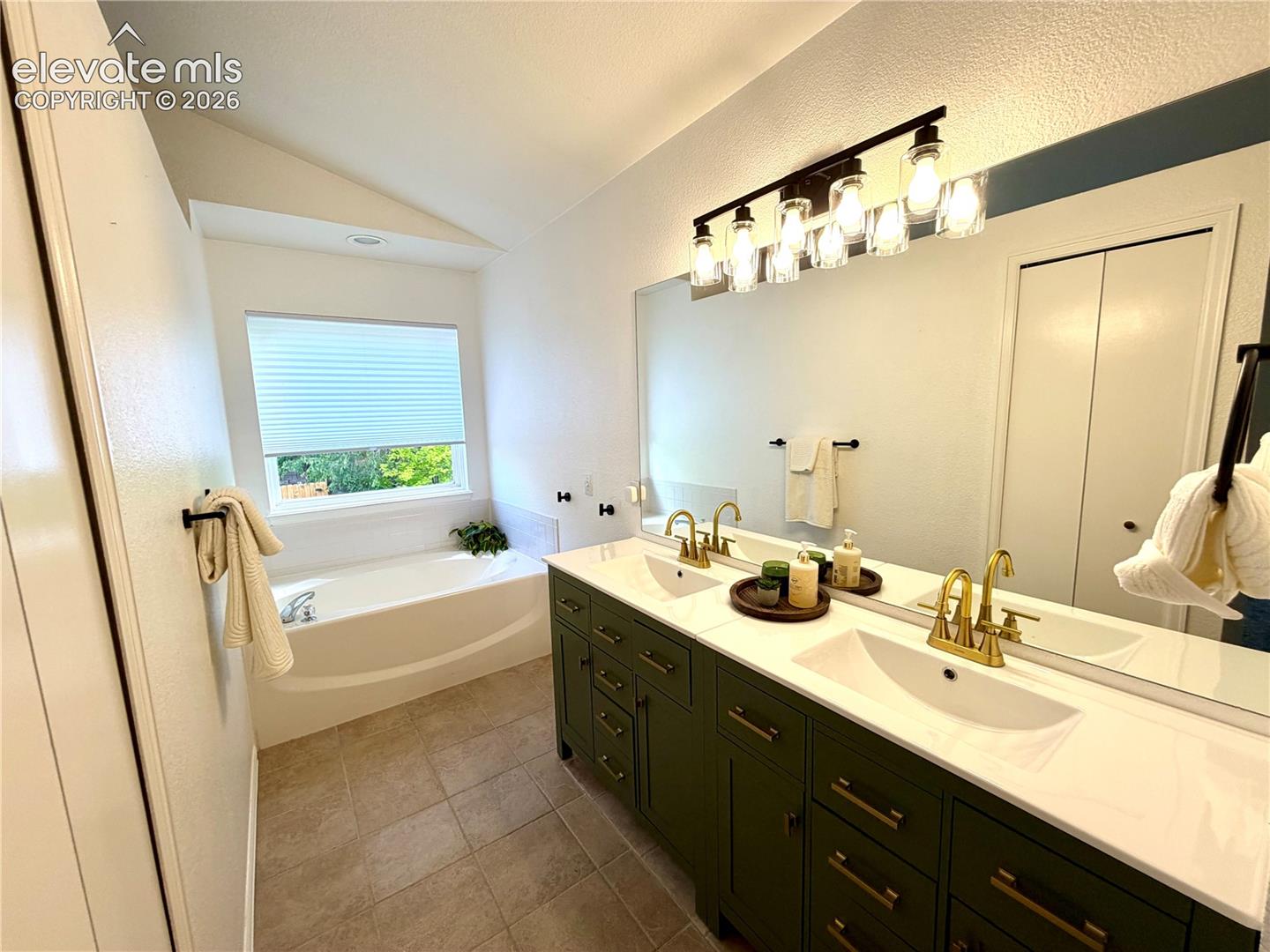 Double vanity with green cabinetry and gold-finish fixtures, a large mirror, a six-light fixture, a built-in tub with a window, and tile flooring