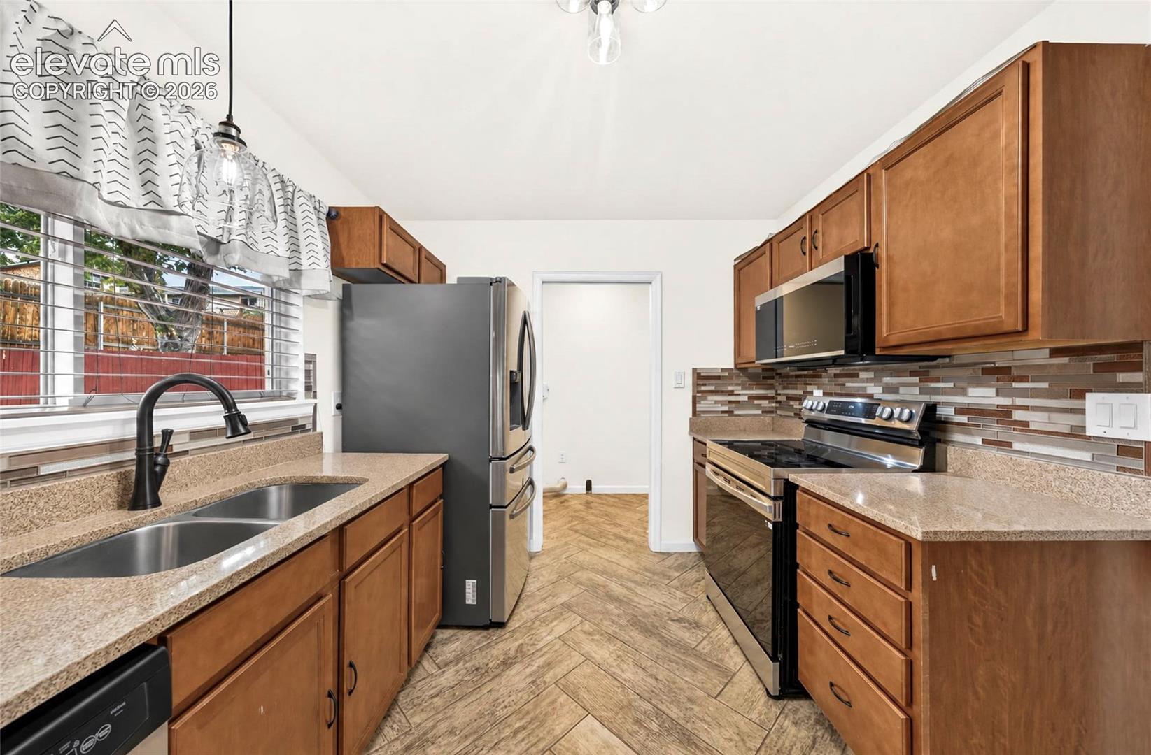 Kitchen featuring wood-finish cabinetry, stone-texture countertops, a multi-tonal tile backsplash, stainless steel appliances, and herringbone-patterned tile flooring