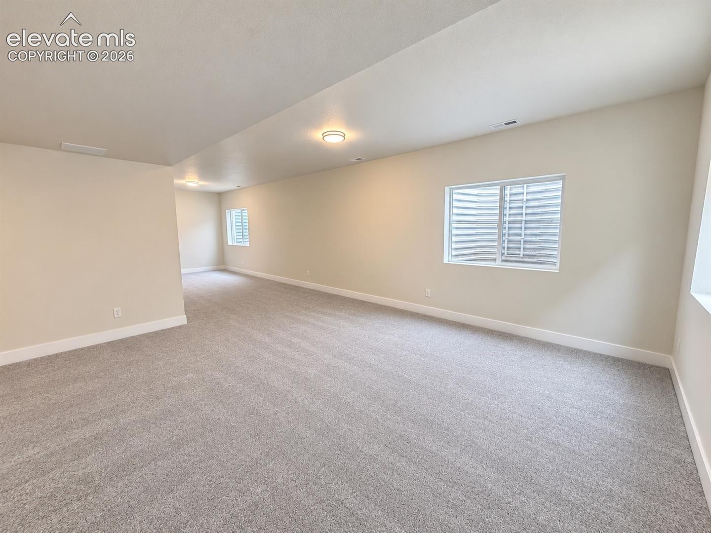 Neutral-toned interior space featuring light-colored wall paint, plush carpeting, ceiling-mounted flush lighting, and multiple egress window wells