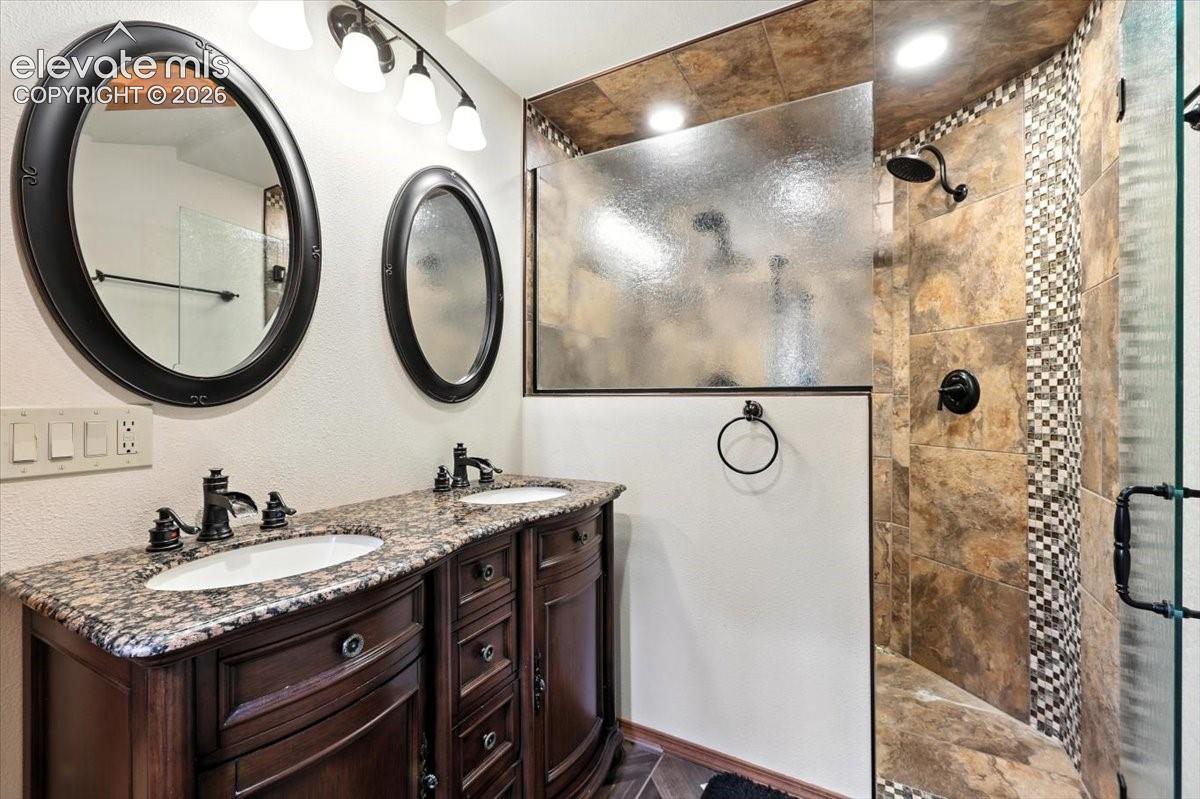 Double-sink vanity with stone-finish countertop, dark wood-finish cabinetry, two oval mirrors with dark frames, and a walk-in shower featuring tile surround and mosaic accent detailing