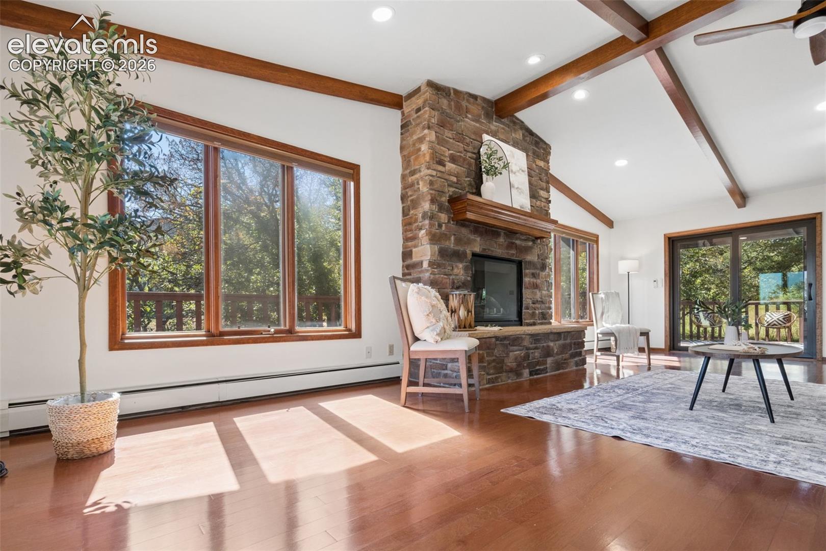 Vaulted ceiling with exposed wood beams, stone fireplace with a wood mantel, wood-finish flooring, recessed lighting, and large windows with wood trim