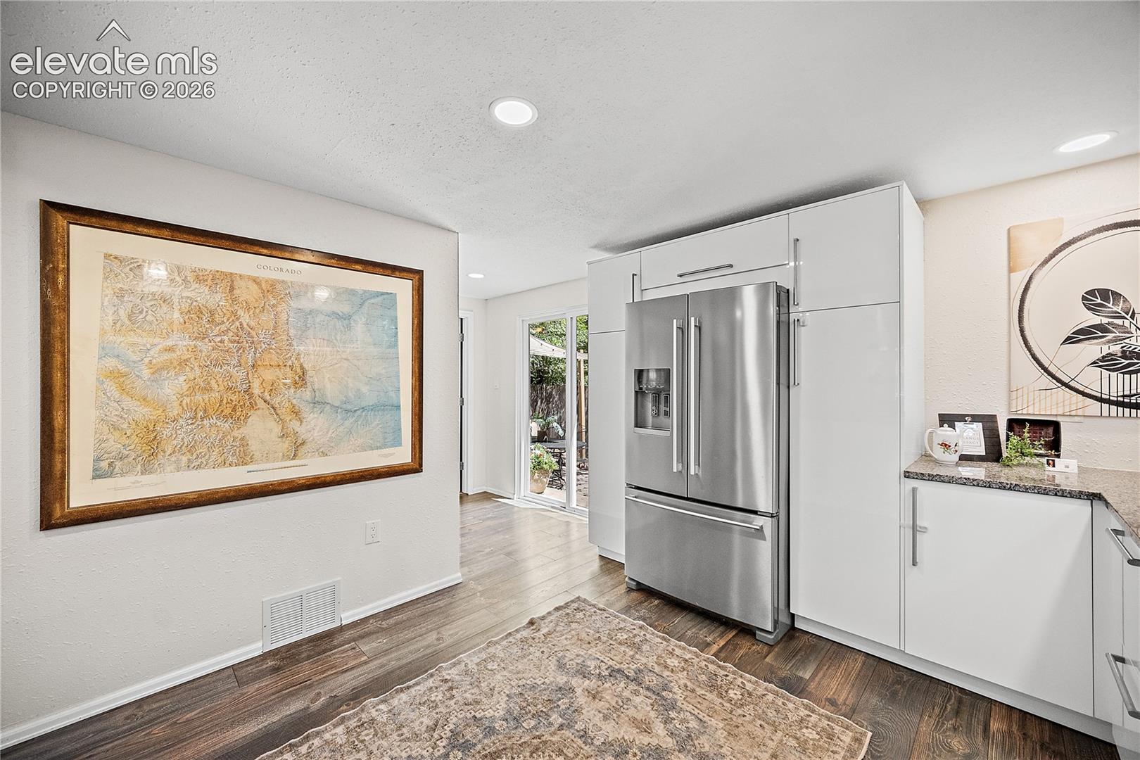 Kitchen area featuring wood-finish flooring, recessed lighting, white cabinetry with silver hardware, a stainless steel refrigerator, and granite countertops