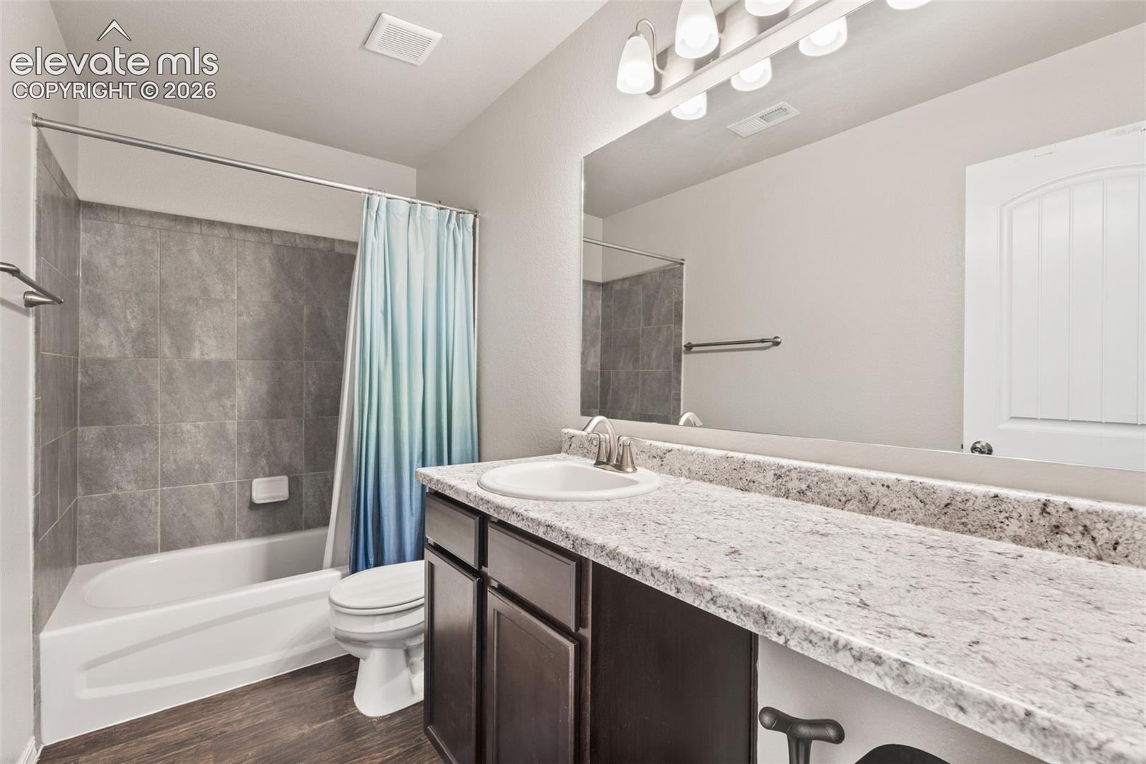 Bathroom featuring a white bathtub, gray tile shower surround, wood-finish flooring, a long vanity with a light-toned countertop, and dark wood-finish cabinetry