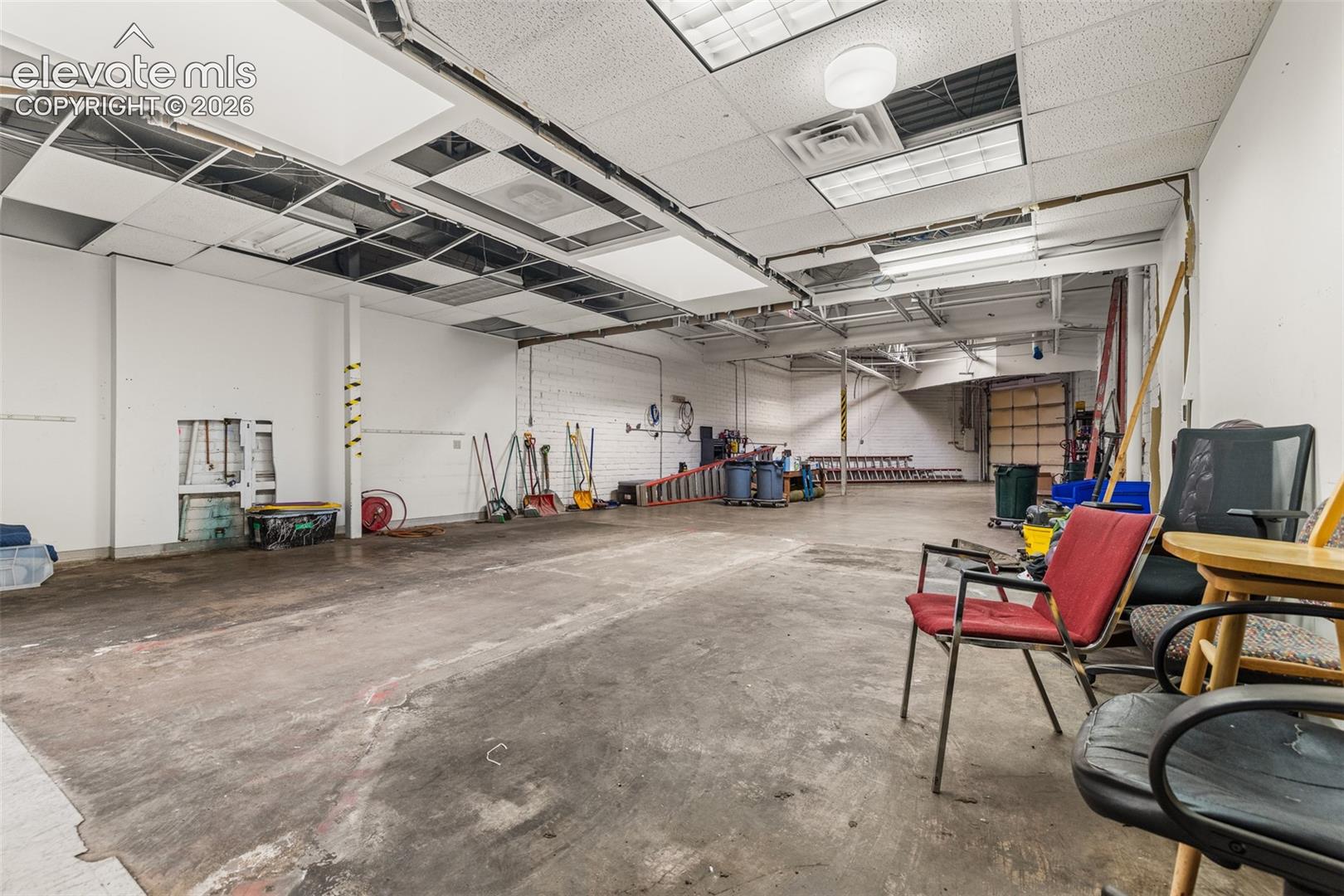 Open-concept commercial space featuring concrete flooring, exposed industrial ceiling grid, drop-ceiling panels, and a roll-up loading door