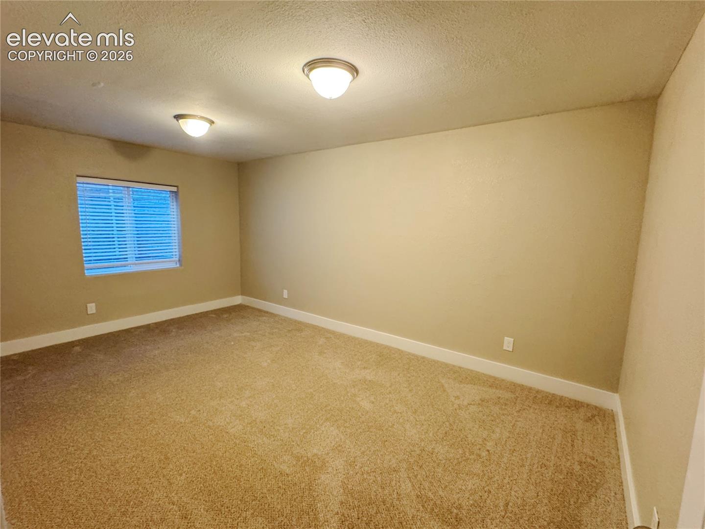 Neutral-toned interior space featuring carpeted flooring, two semi-flush mount light fixtures, and a window with horizontal blinds