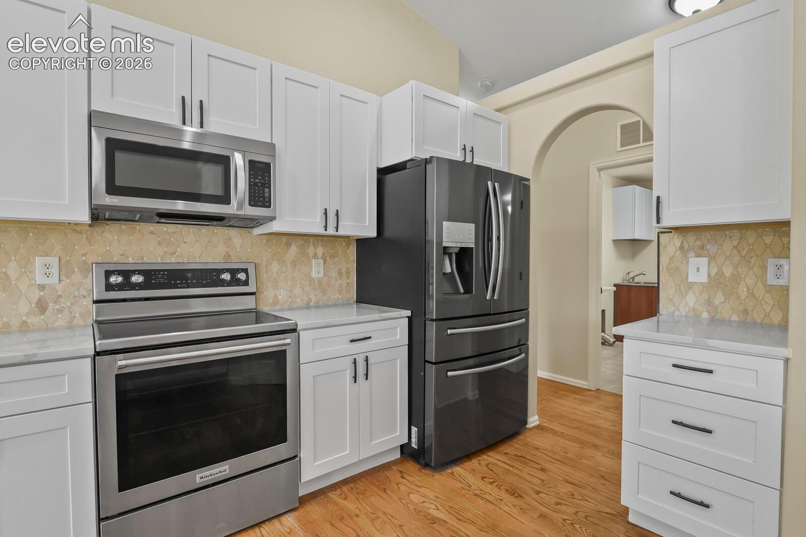 Modern kitchen featuring stainless steel appliances, white cabinetry with dark hardware, light-toned backsplash, and light wood-finish flooring