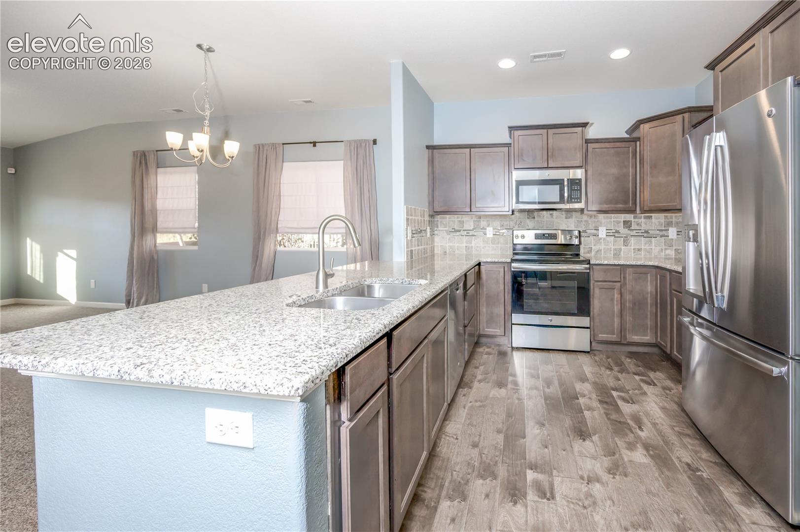 Kitchen featuring a stone-composite countertop island, stainless steel appliances, wood-finish flooring, shaker-style cabinetry, and a multi-toned tile backsplash