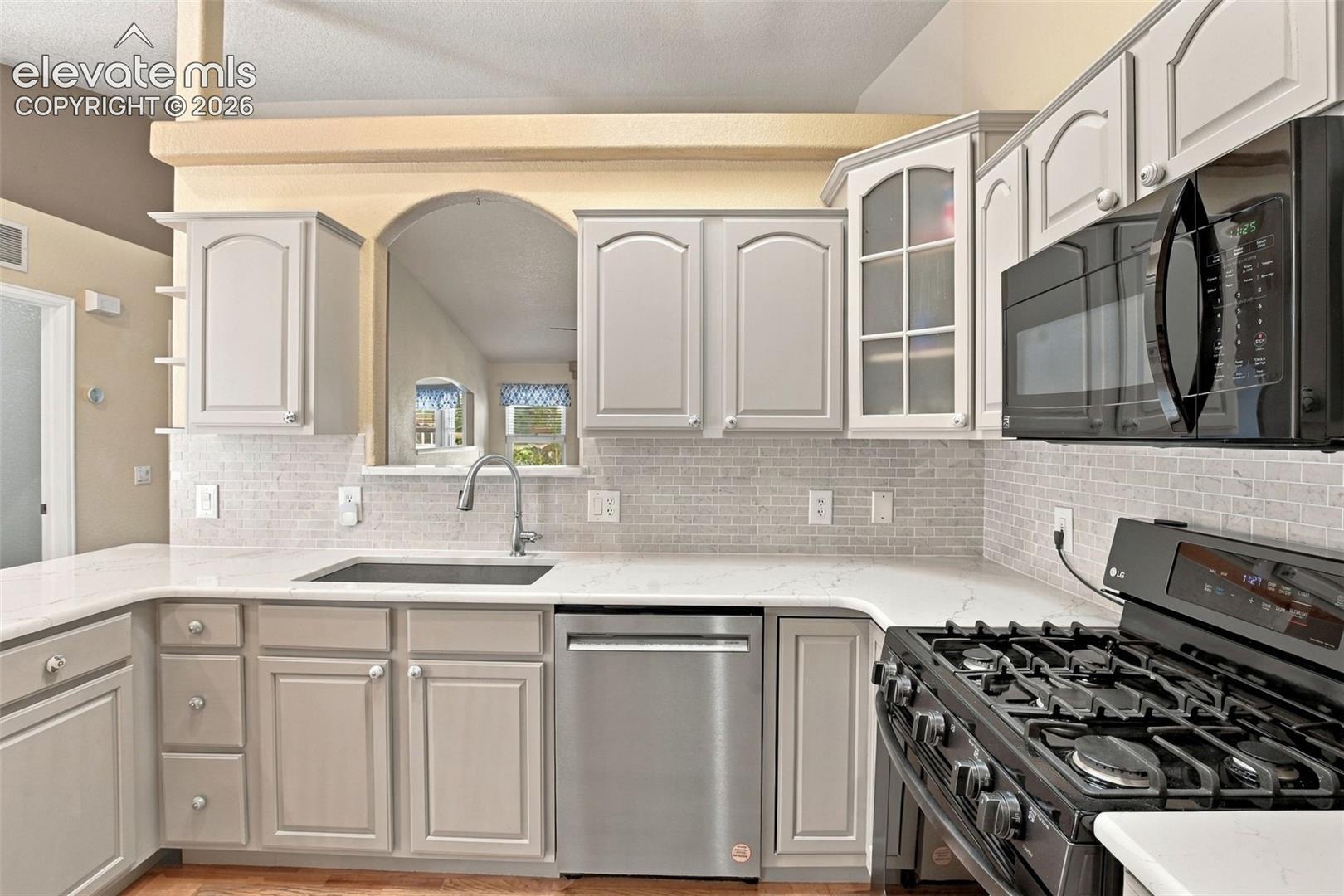 Kitchen featuring light-toned cabinetry, solid surface countertops, a decorative tile backsplash, stainless steel dishwasher, and an arched pass-through window