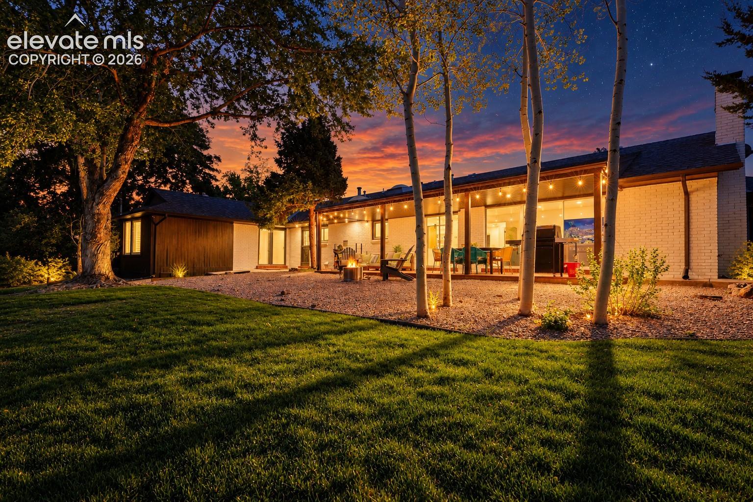 Spacious outdoor patio with string lighting, expansive lawn, mature trees, and a fire pit