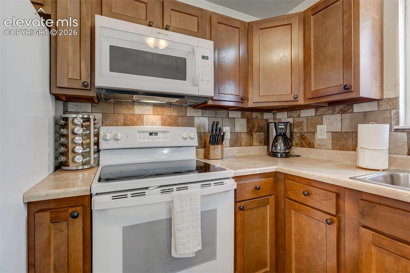 Wood-finish cabinetry, solid surface countertops, tile backsplash, stainless steel sink, and a white oven with an overhead microwave