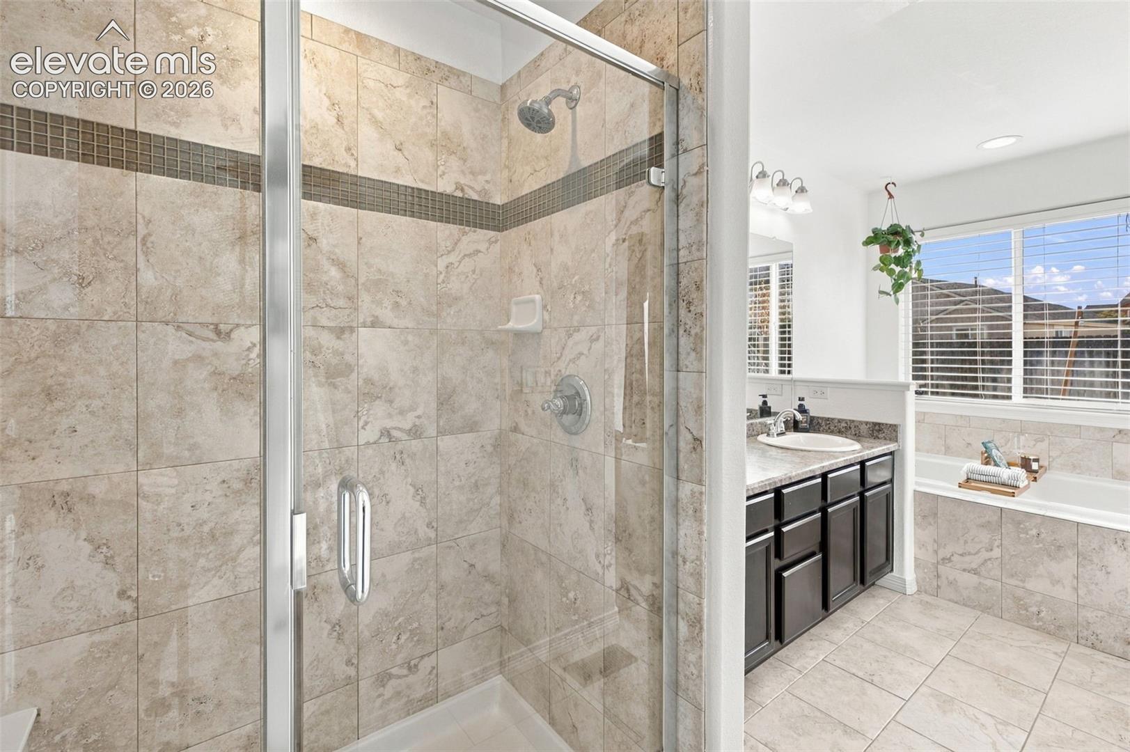 Walk-in glass-enclosed shower featuring stone-finish wall tile with a mosaic accent border, dark wood cabinetry with stone-surface countertops, dual-basin vanity, and a drop-in bathtub