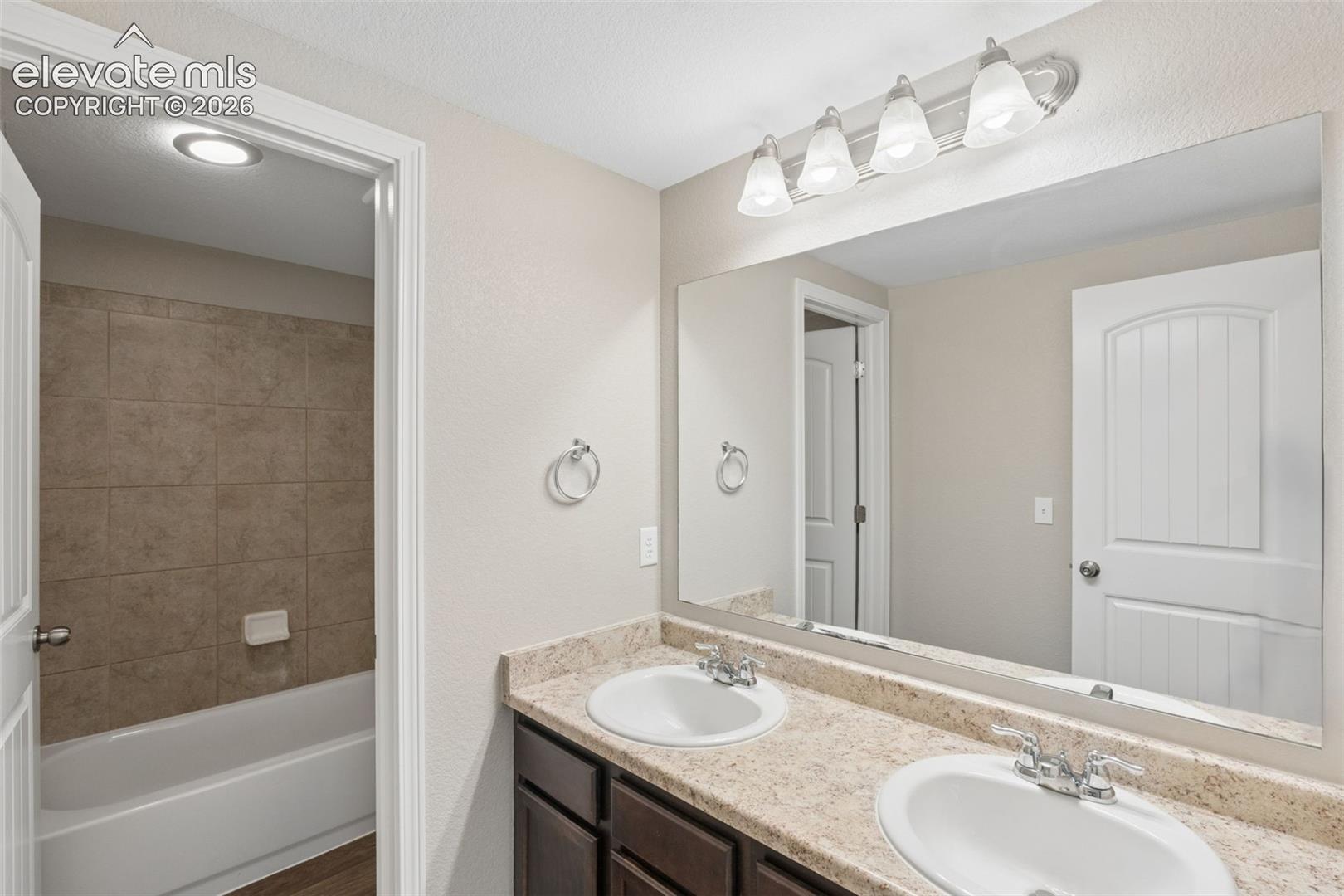 Bathroom in basement featuring a dual sink vanity with a speckled countertop, a large frameless mirror, and a four-light fixture