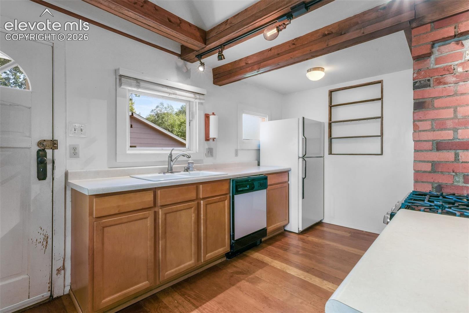 Kitchen featuring wood-finish flooring, exposed wood ceiling beams, a window over the sink, and a brick accent wall