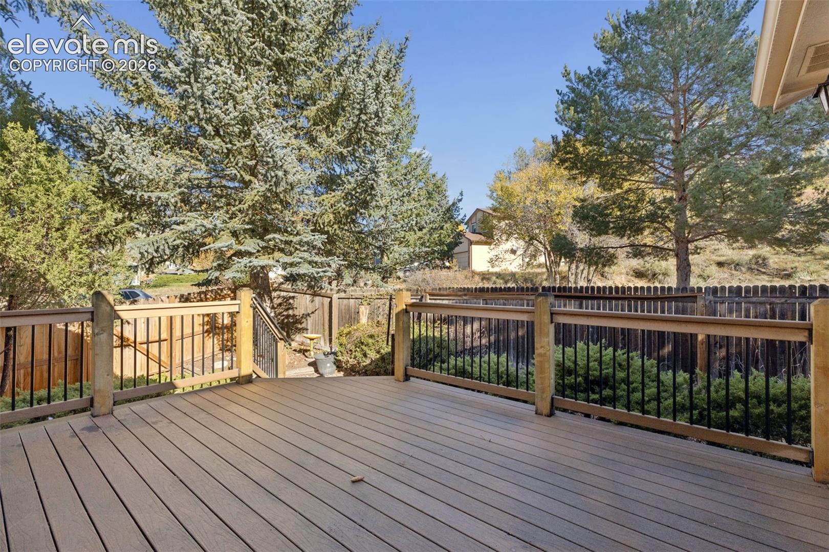 Raised wood-finish deck with black metal railing, mature evergreen trees, and wood privacy fencing