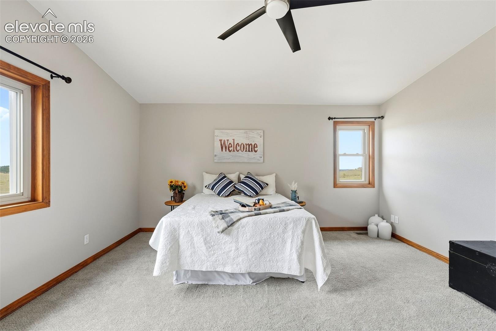 Vaulted ceiling, two windows with wood-trim, light gray carpet, and a ceiling fan