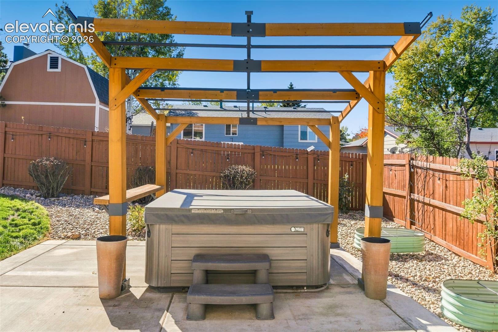 Custom wooden pergola structure situated over a built-in hot tub on a concrete patio, enclosed by a wood privacy fence and decorative rock landscaping