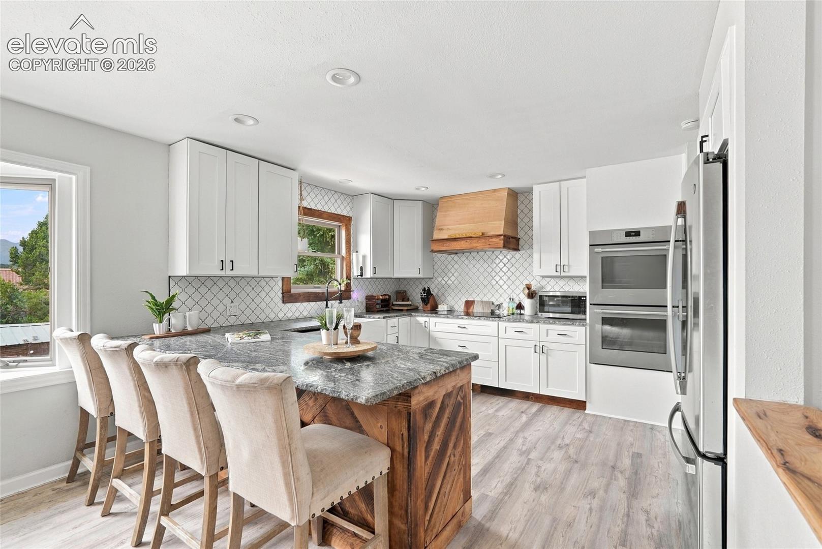 White cabinetry with contemporary hardware, stainless steel double wall ovens, wood-finish flooring, a kitchen island with a stone countertop, and a wood range hood