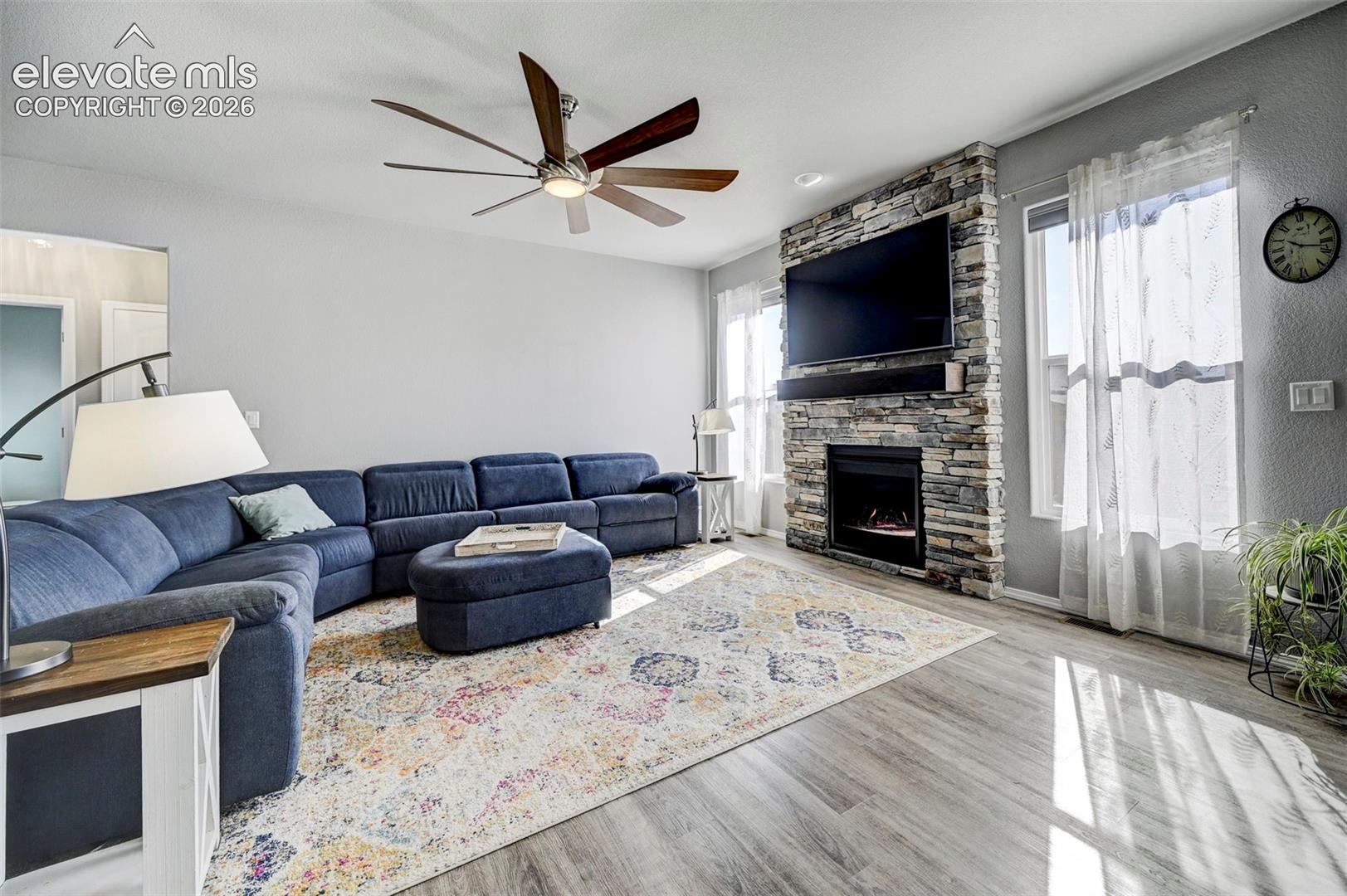 Living area featuring a floor-to-ceiling stacked stone fireplace with an integrated mantel, a multi-blade ceiling fan, light gray walls, and wood-finish flooring