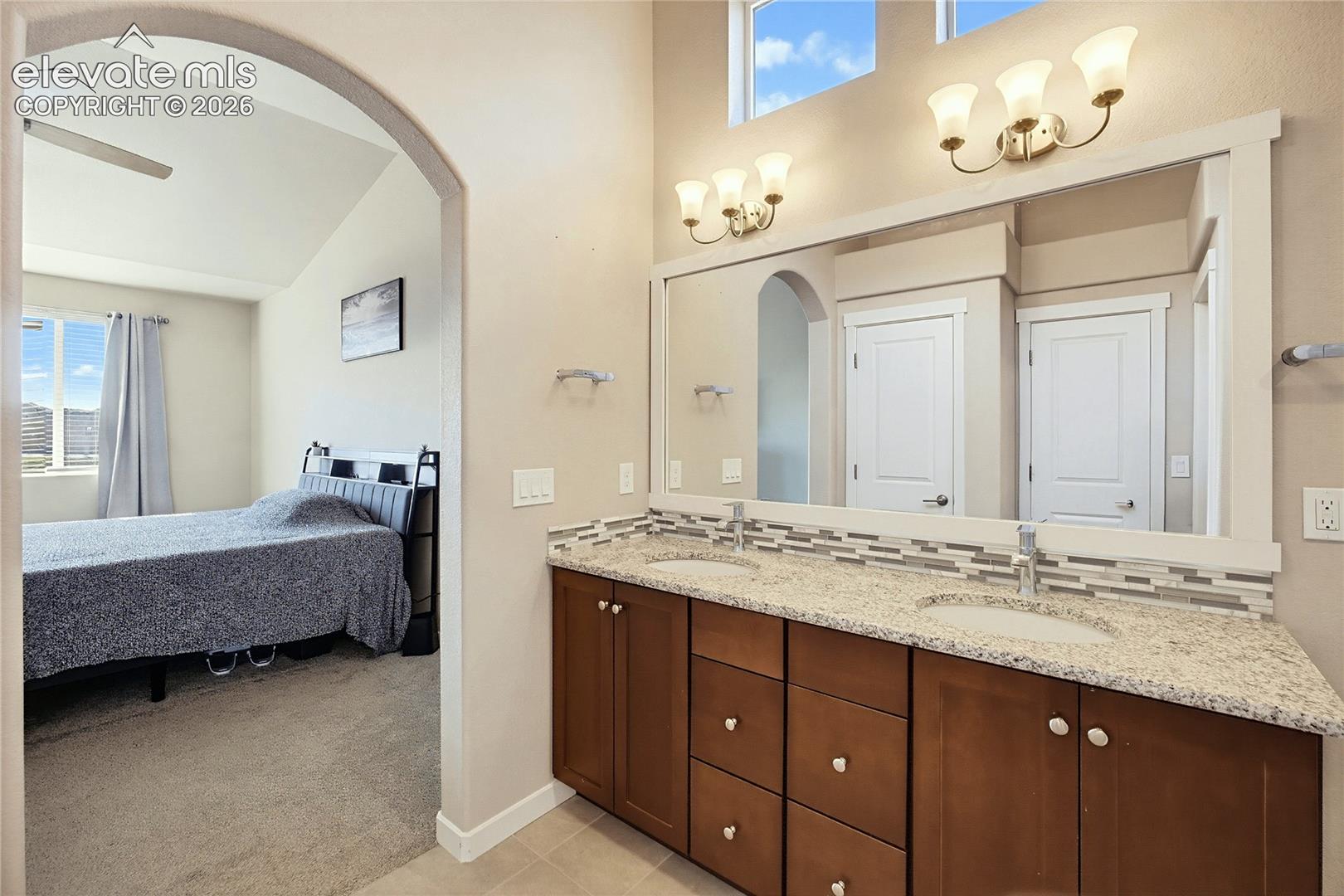 Double vanity with stone countertop, mosaic tile backsplash, framed wall mirror, and dual three-light wall fixtures