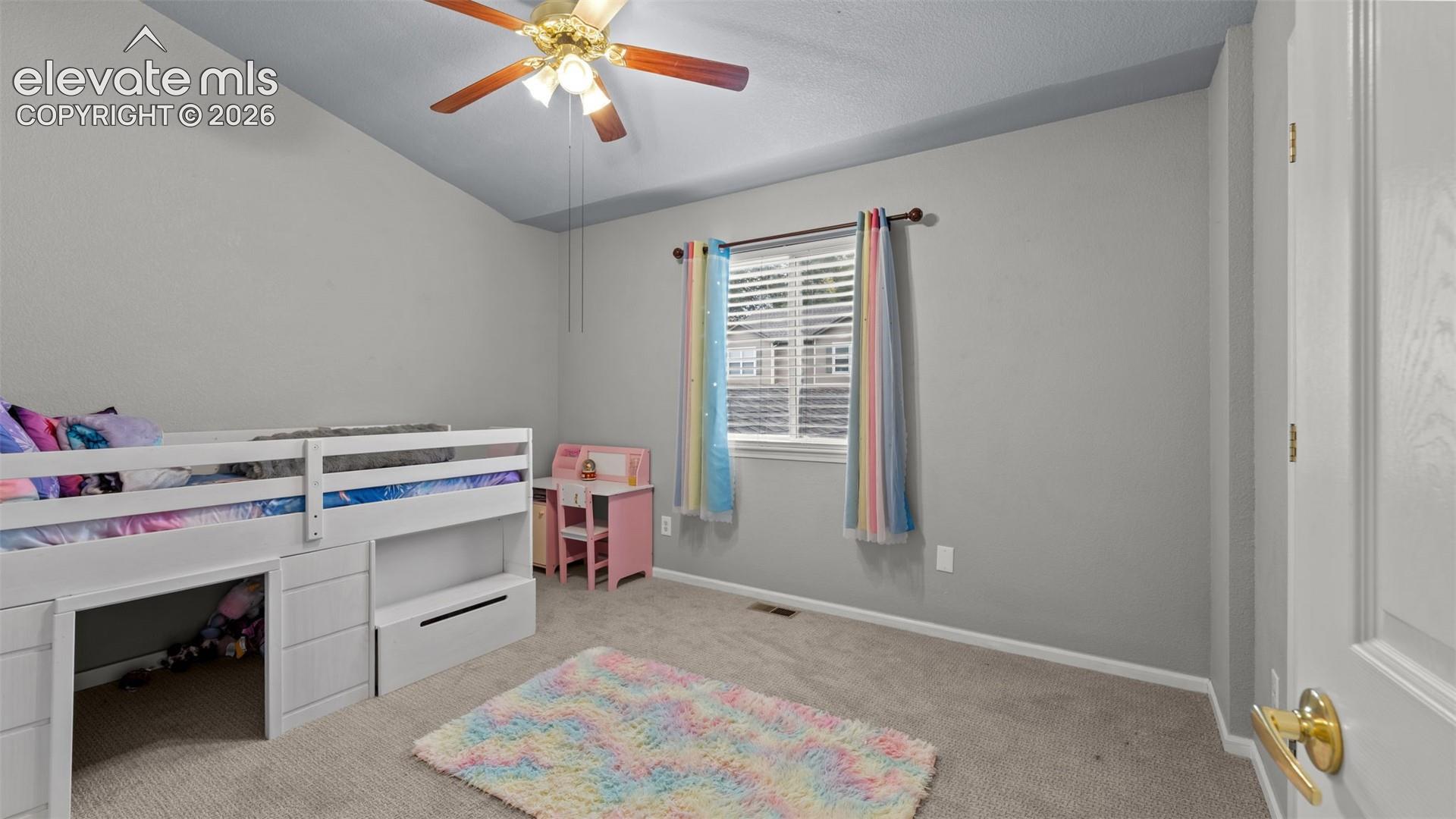 Vaulted ceiling featuring a central ceiling fan with multi-blade finish and light kit, neutral-tone carpeting, and a window with horizontal blinds