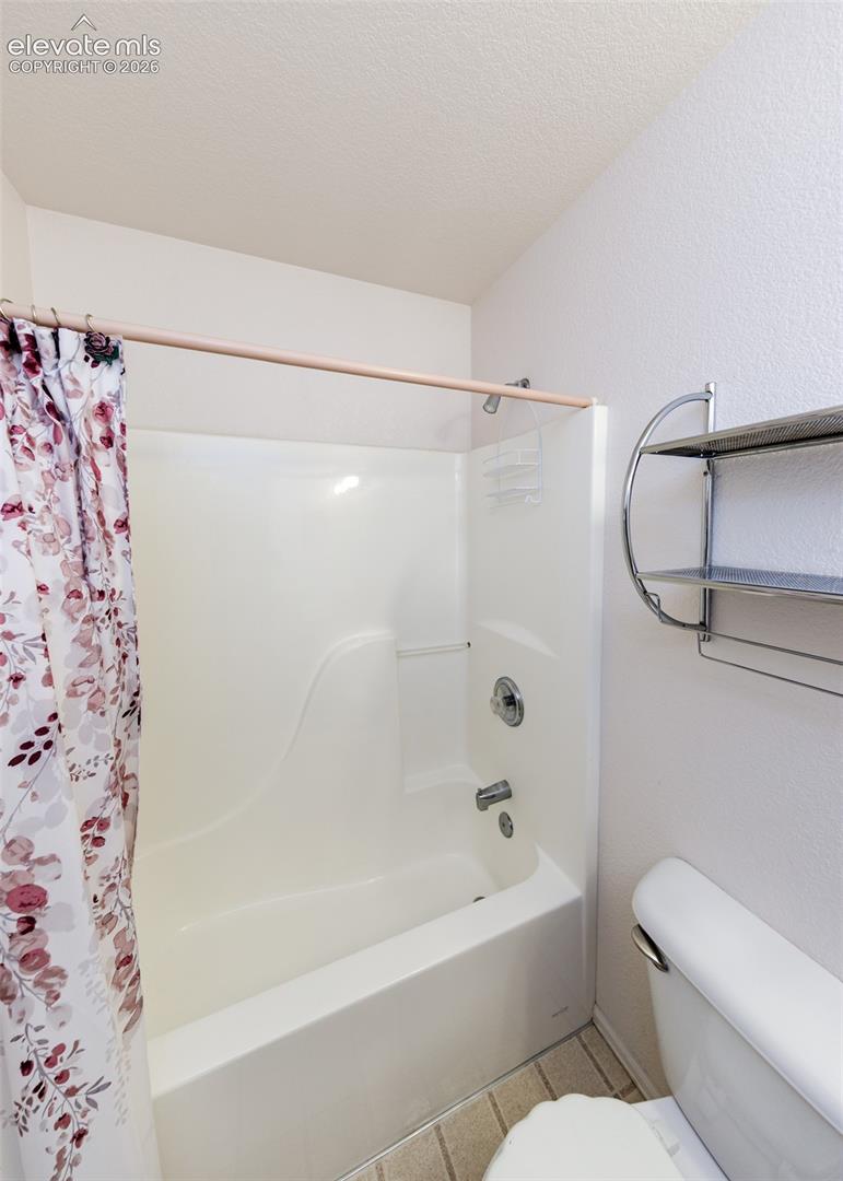 White molded bathtub and wall surround featuring a chrome-finish shower head, mixing valve, and spout, complemented by wall-mounted metal shelving and tile flooring