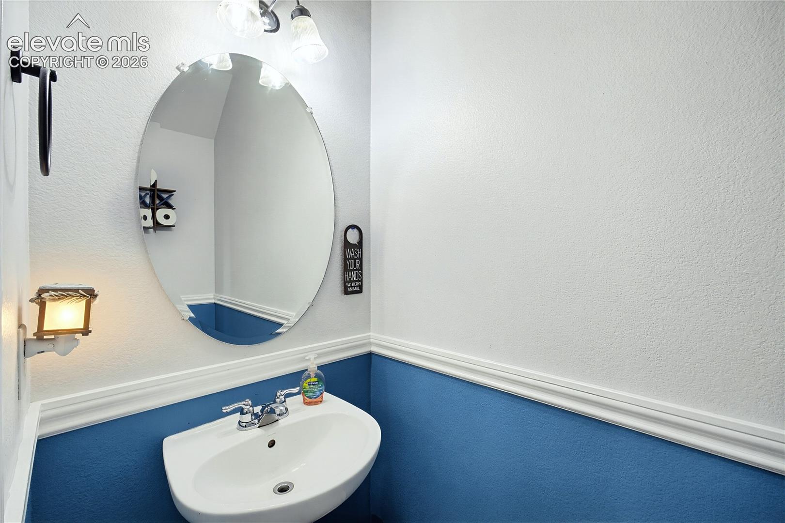 Wall-mounted sink with chrome faucet, an oval frameless mirror, and a three-light vanity fixture