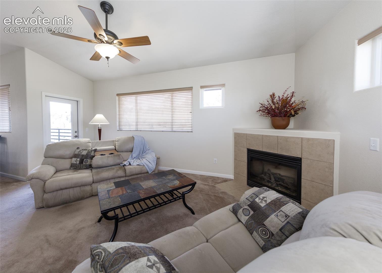 Fireplace with tile surround, vaulted ceiling, ceiling fan with integrated lighting, and horizontal window blinds