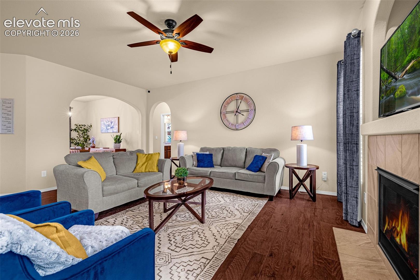 Wood-finish flooring, fireplace with tile surround, arched architectural openings, and ceiling fan with integrated light fixture
