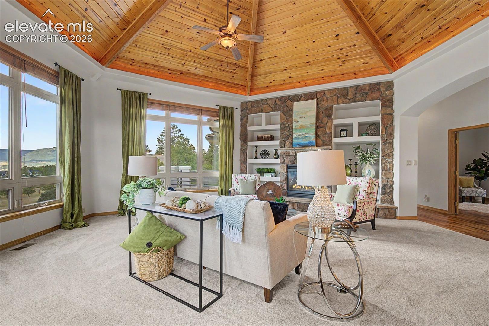 Living area featuring a vaulted wood-paneled ceiling with exposed beams, a stone-faced fireplace with built-in shelving, expansive windows, and light-toned carpeting