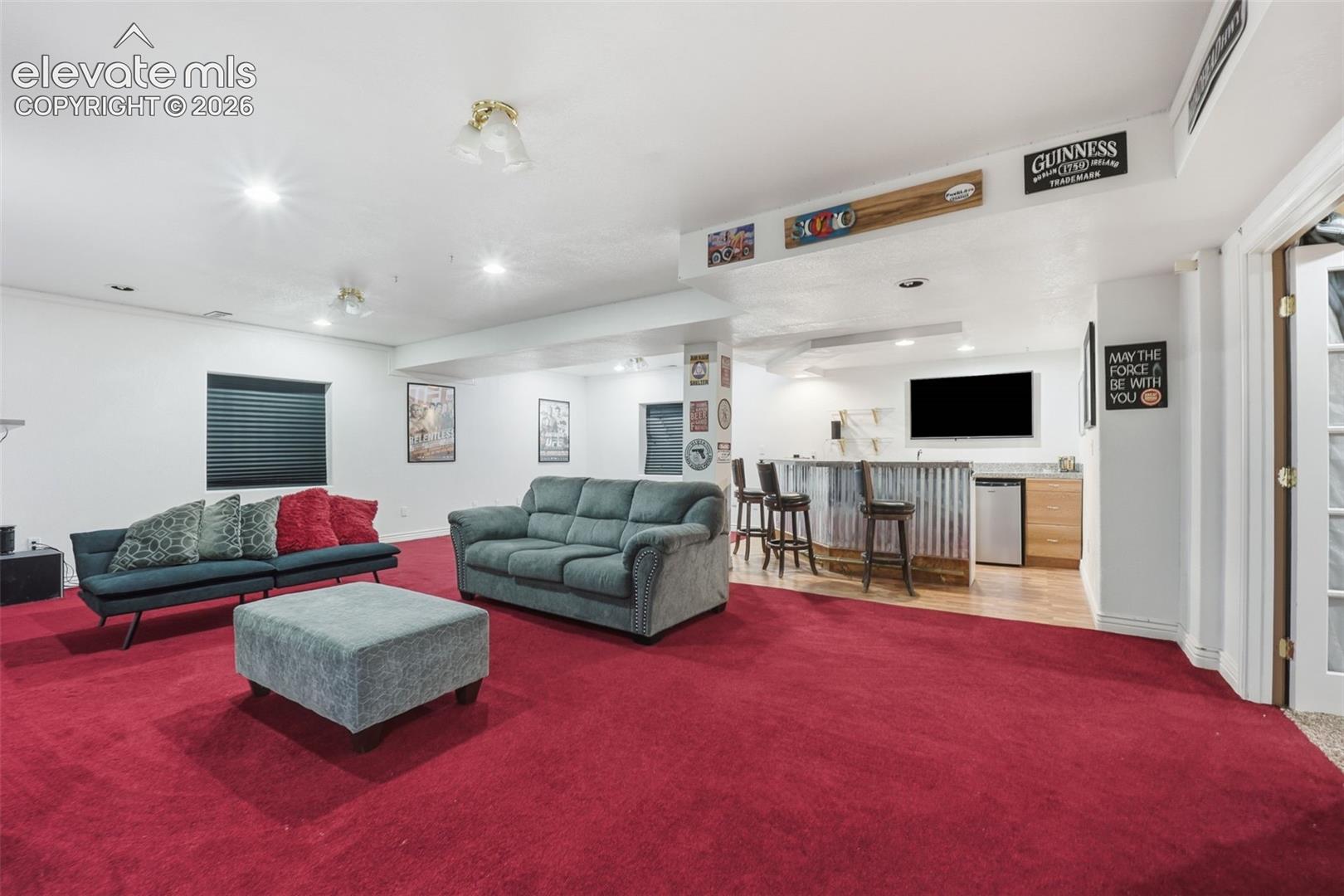 Spacious finished basement featuring recessed lighting, a built-in bar with a corrugated metal facade, wood-finish flooring at the bar area, and a mini-refrigerator