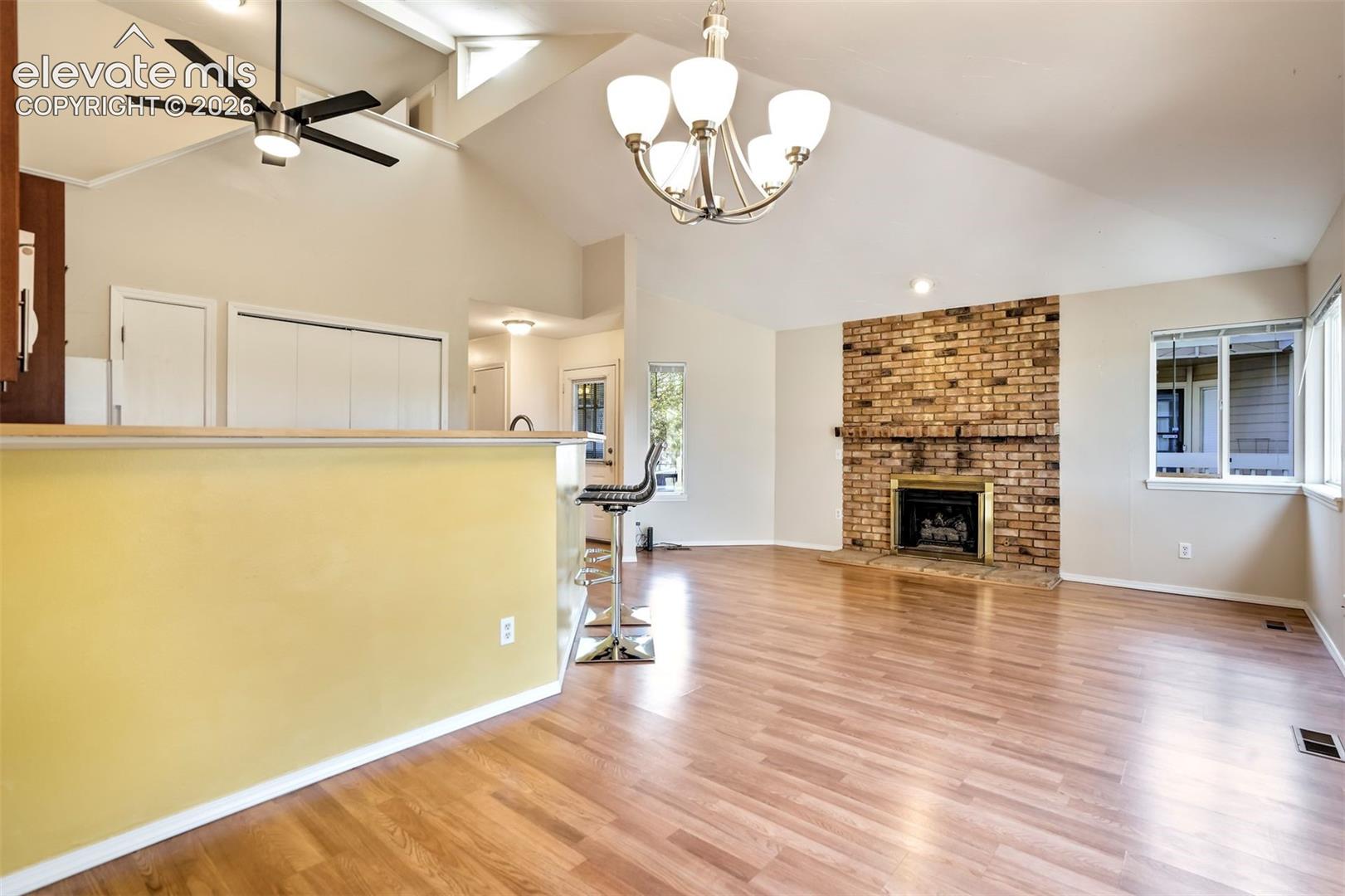 Vaulted ceilings, brick fireplace with a metallic surround, wood-finish flooring, multi-arm chandelier, and a breakfast bar with a solid surface countertop