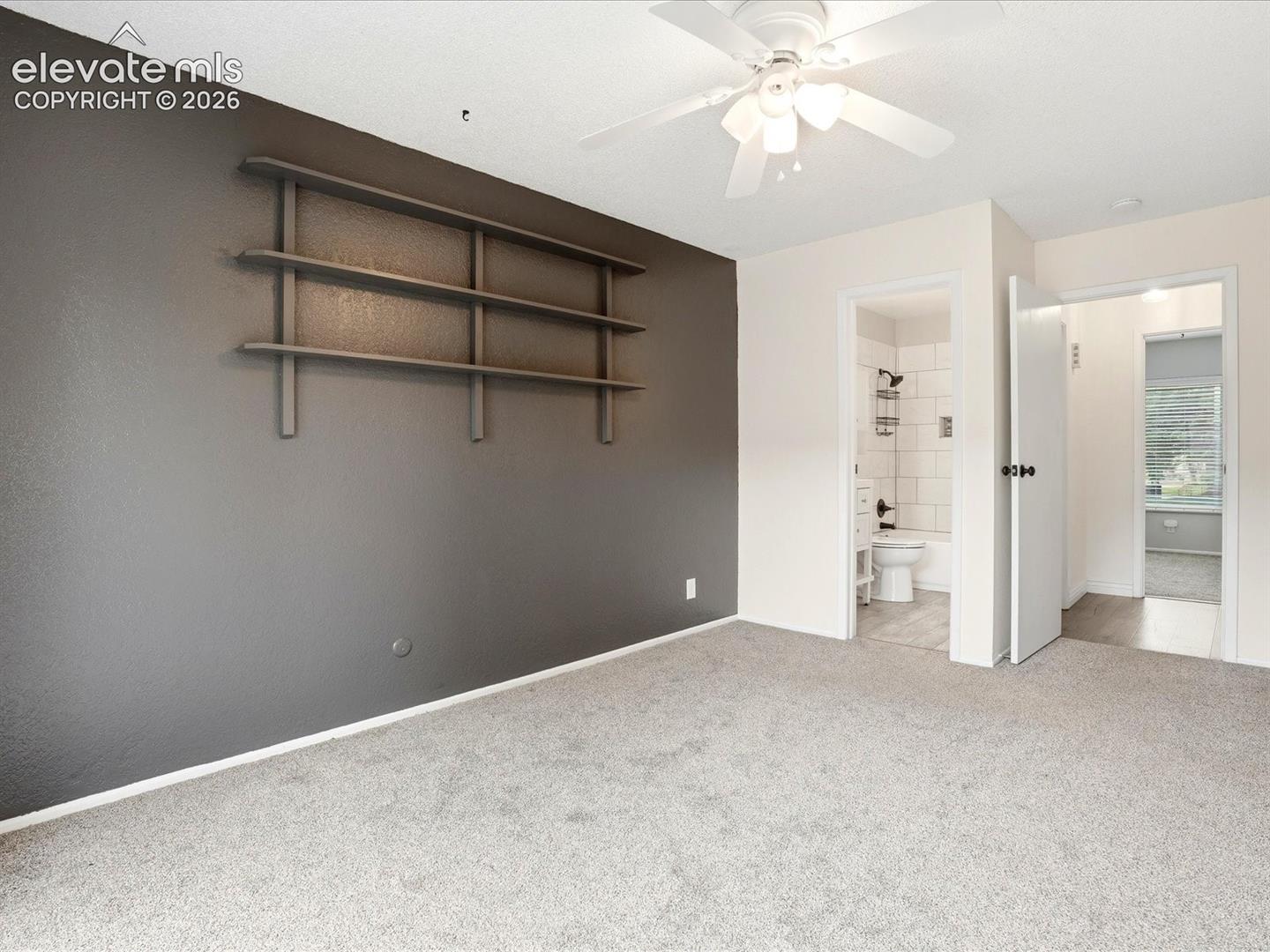 Room featuring a dark-toned accent wall with built-in shelving, neutral wall-to-wall carpeting, and a ceiling fan with integrated lighting