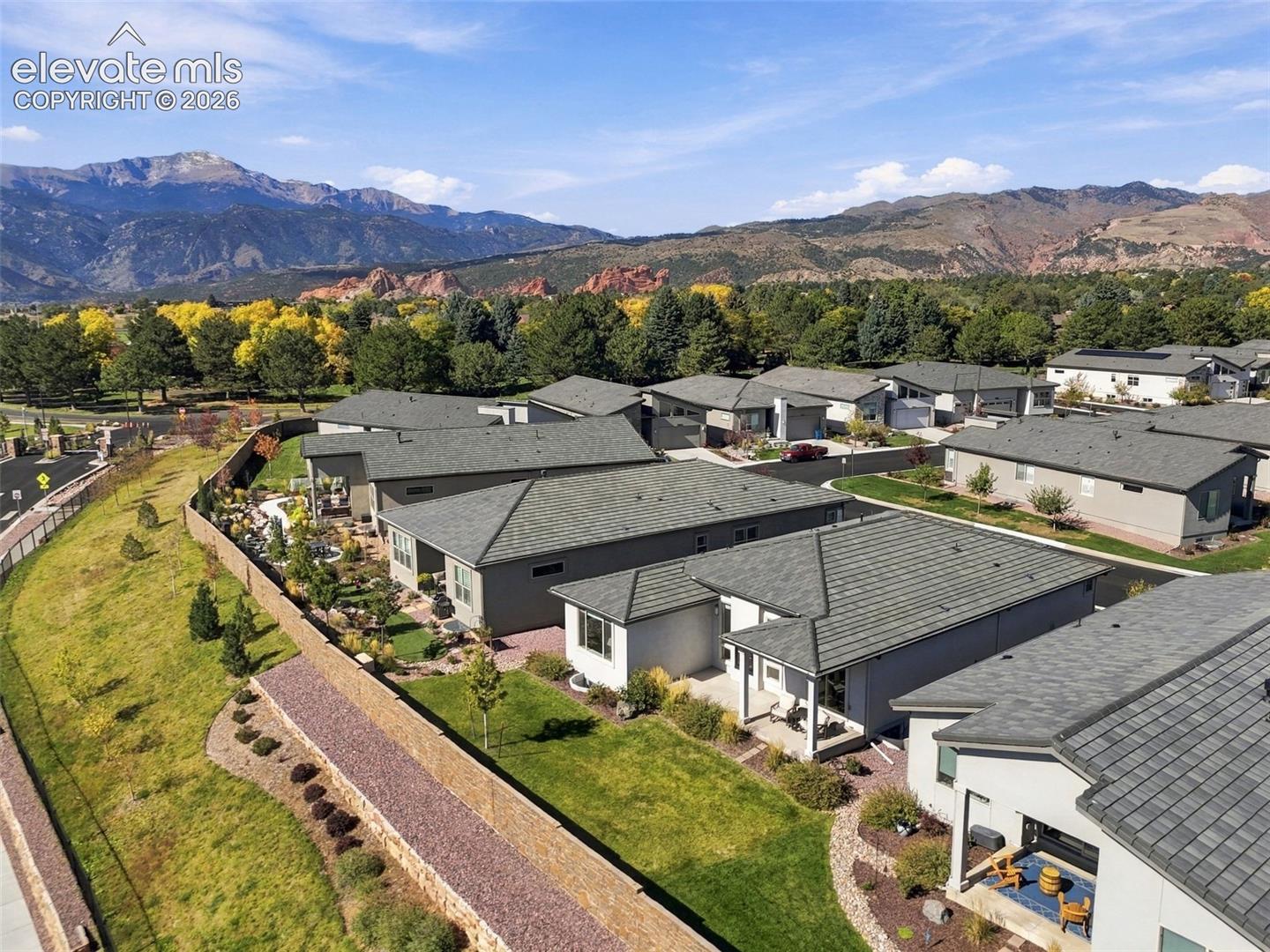 Aerial perspective featuring residential structures with asphalt shingle roofs, exterior stucco siding, manicured lawns, stone retaining walls, and a prominent mountain range backdrop