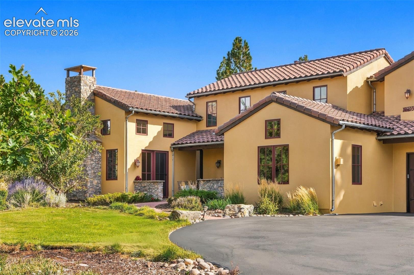 Stucco exterior with a tiled roof, stone chimney, paved driveway, and manicured landscaping