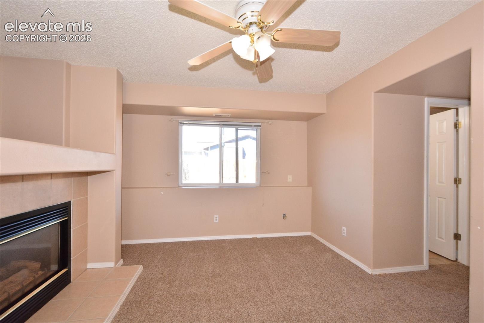 Carpeted room featuring a built-in fireplace with a tiled hearth and surround, a ceiling fan with light fixture, and a window