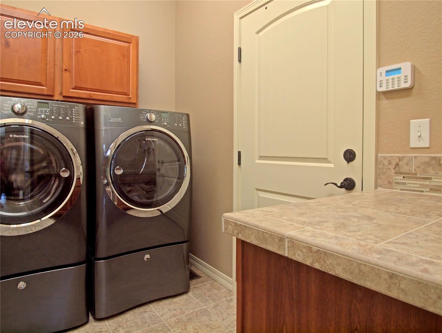 Dedicated laundry area, storage closet  and garage door exit, featuring a tiled countertop, wood-finish cabinetry, and a decorative tile backsplash