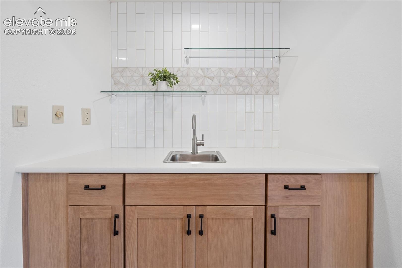 Wood-finish cabinetry with black hardware, white countertop, stainless steel sink with a gooseneck faucet, white subway tile backsplash, and decorative geometric tile accent