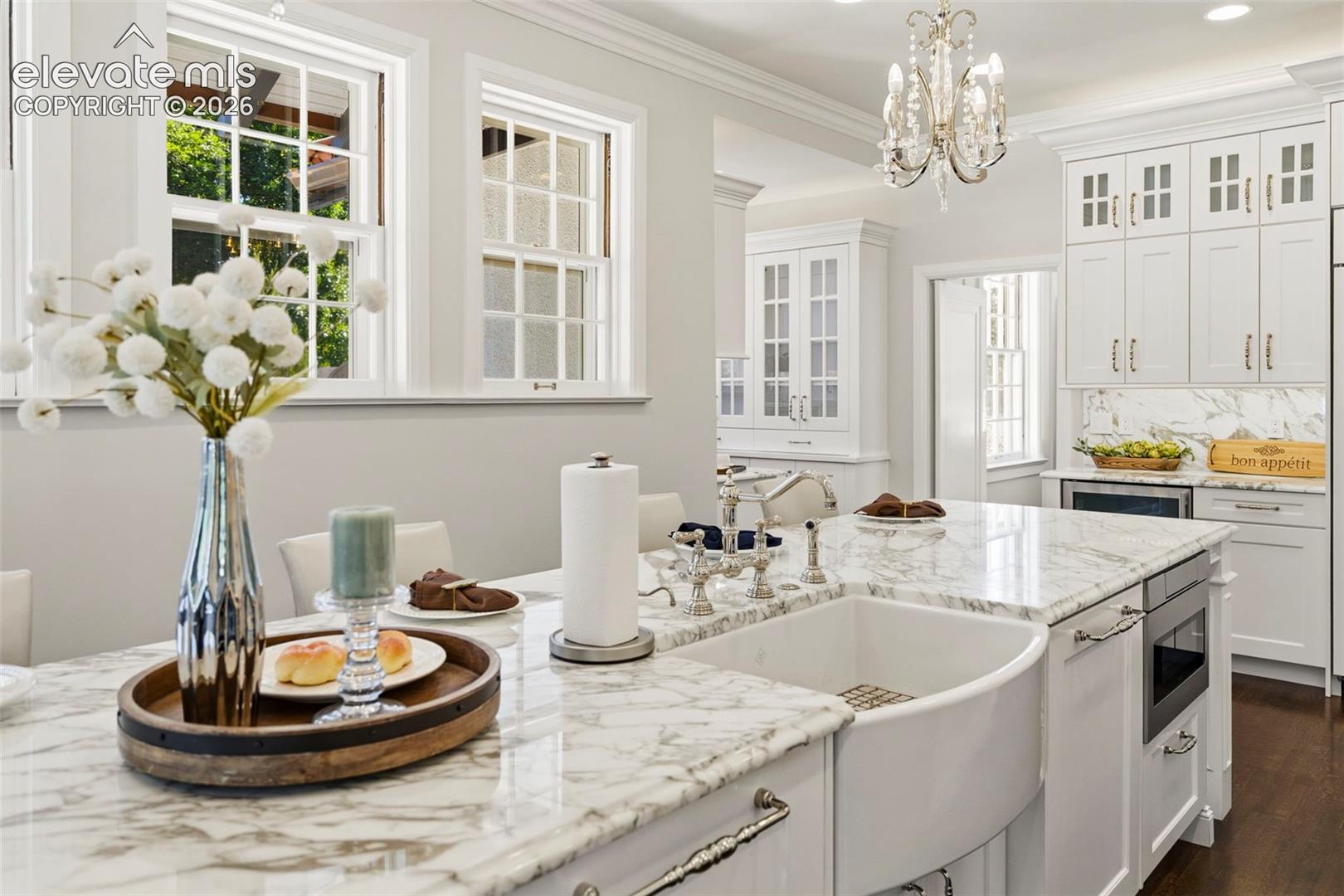 Kitchen featuring white cabinetry with glass-front uppers, stone countertops, a farmhouse-style apron sink, polished metal hardware, and a tiered crystal chandelier