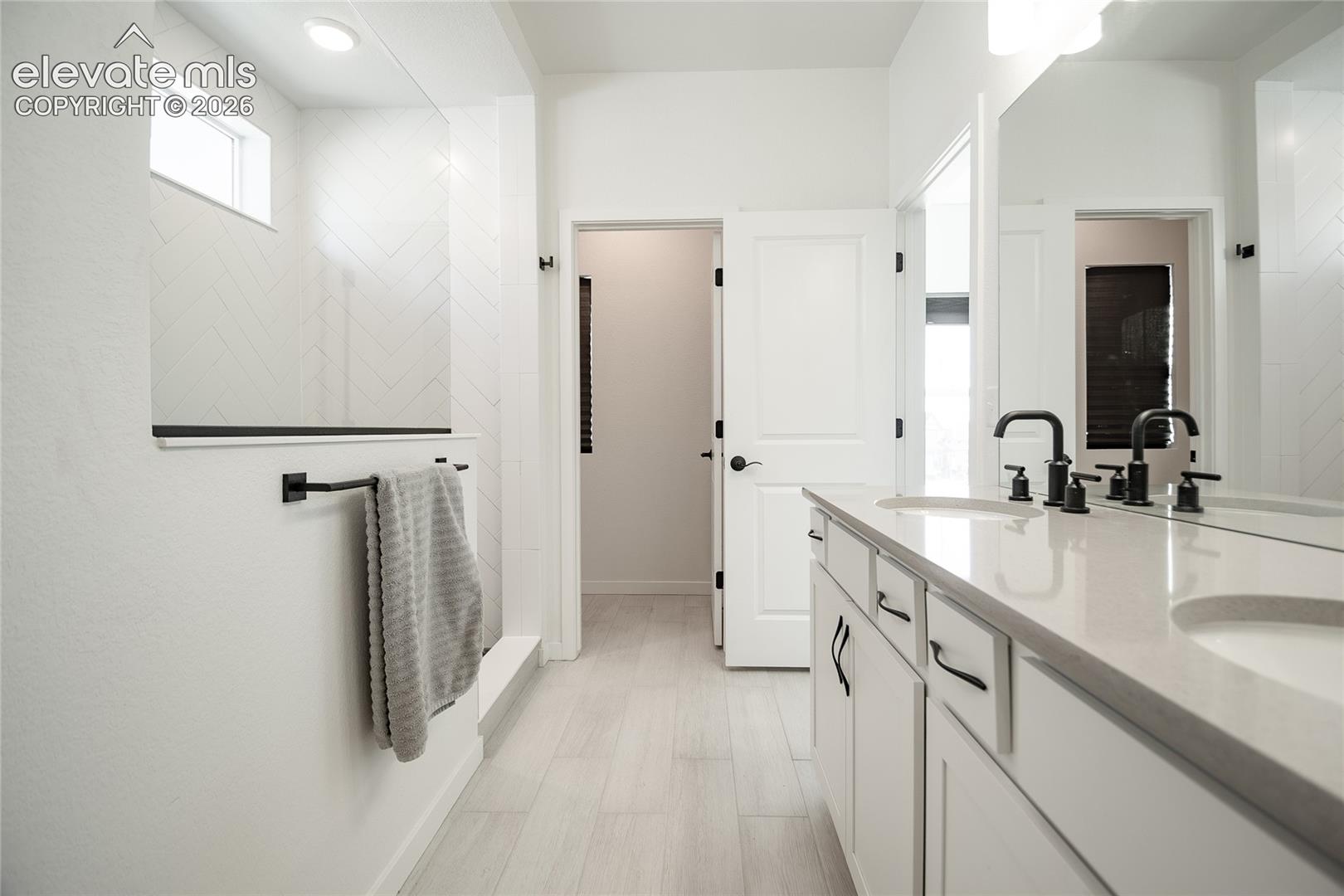 Dual-sink vanity with stone-finish countertop, matte black fixtures, herringbone tile shower surround, and light wood-finish flooring