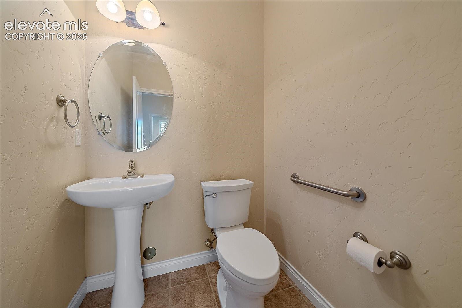 Powder room featuring a white pedestal sink, oval wall mirror, dual-light fixture, tile flooring, and a stainless steel grab bar
