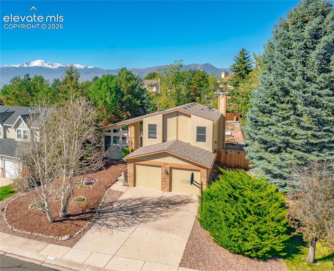 Two-car garage with basketball hoop, concrete driveway, front yard landscaping with mature trees, and snow-capped mountain backdrop