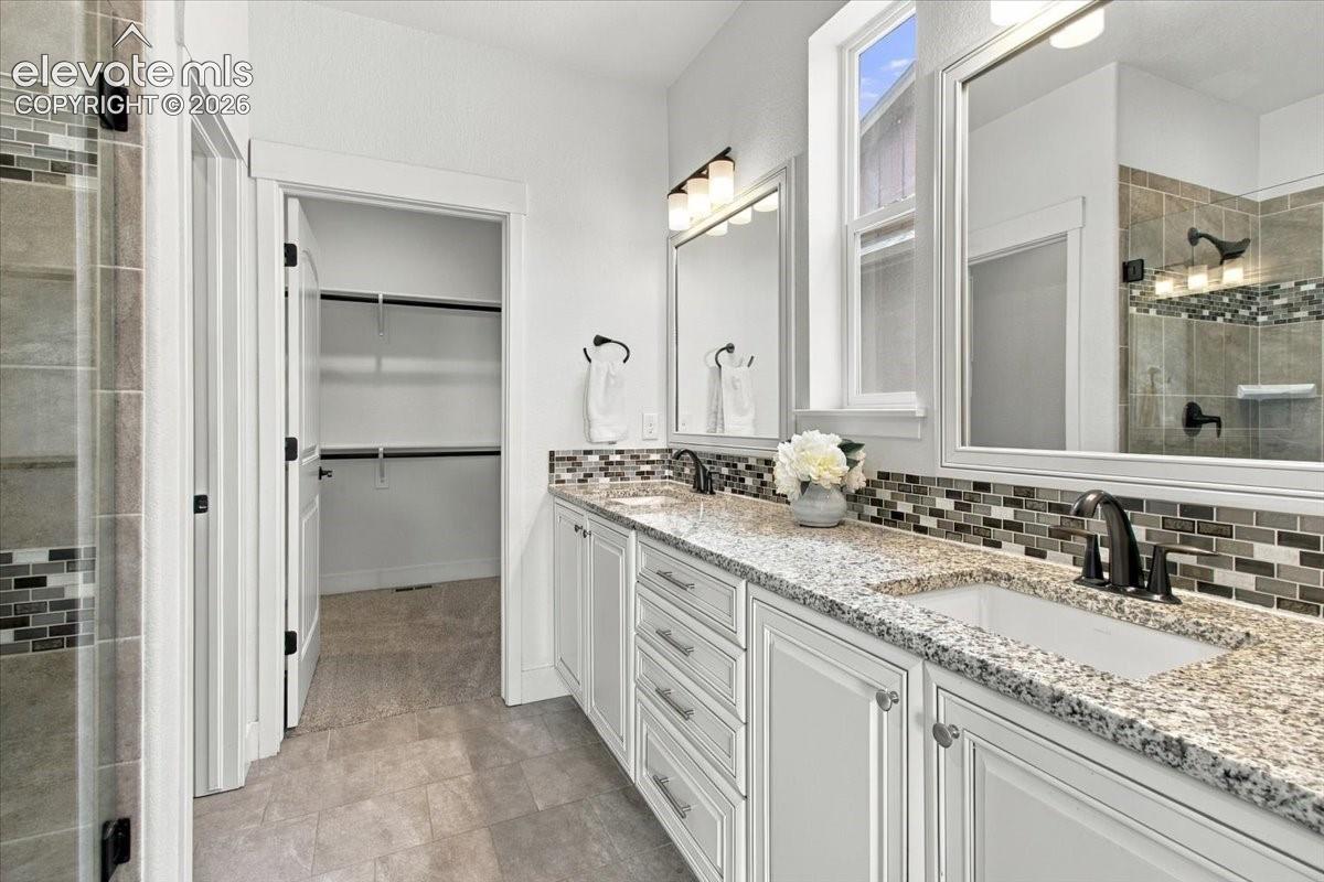Double vanity featuring white cabinetry, stone-finish countertops, undermount sinks, and a multi-tone tile backsplash, paired with a walk-in closet featuring built-in shelving