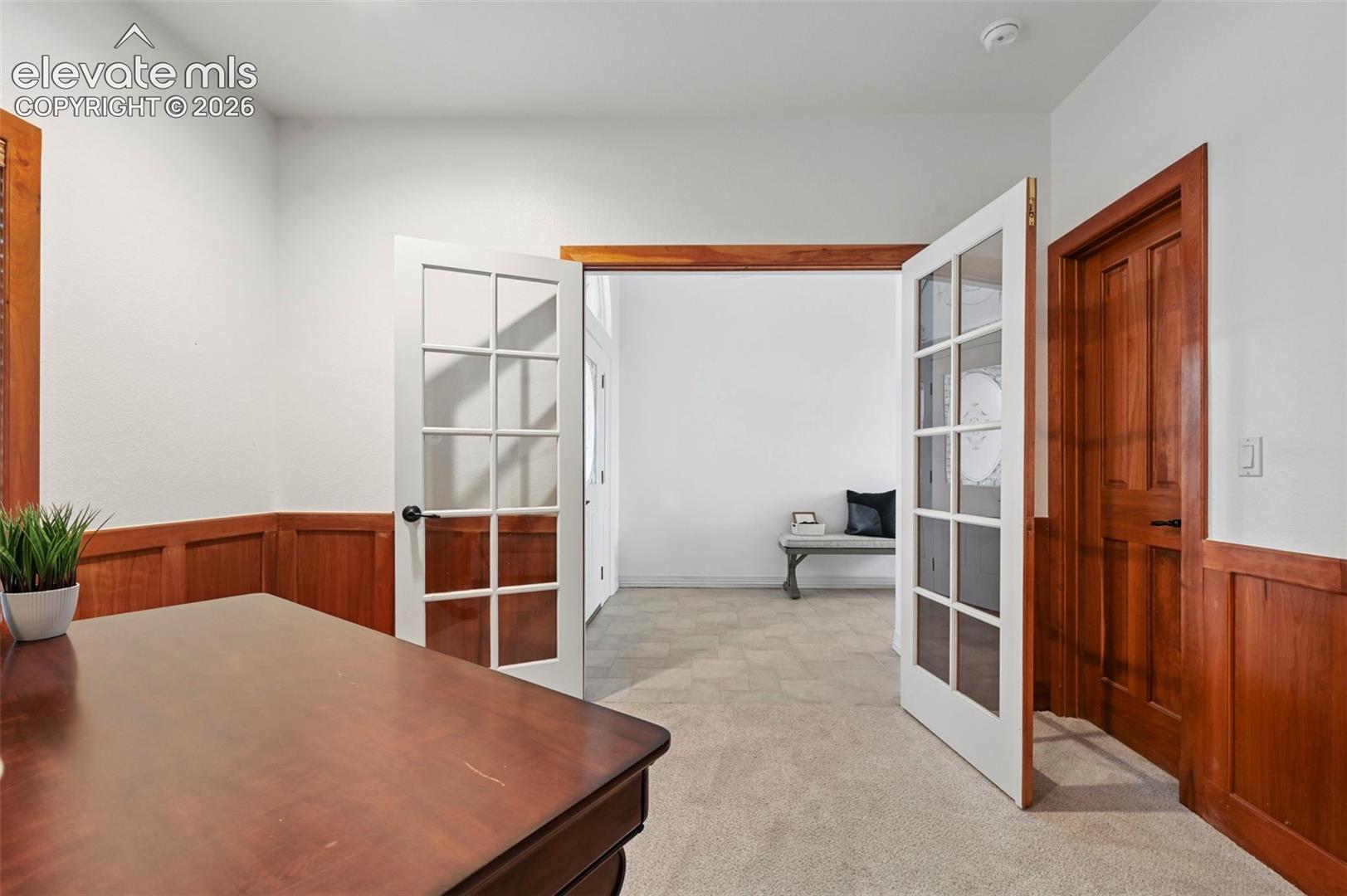 Interior room featuring white walls with wood wainscoting, a light-toned carpet, and a pair of white French doors with divided lites