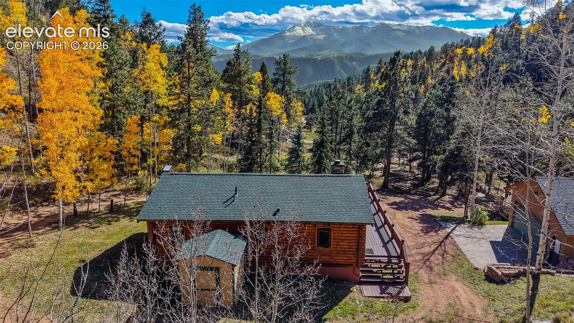Log cabin exterior featuring a shingle roof, chimney, expansive wood deck, and detached storage structure, set against a mountainous backdrop with mature trees