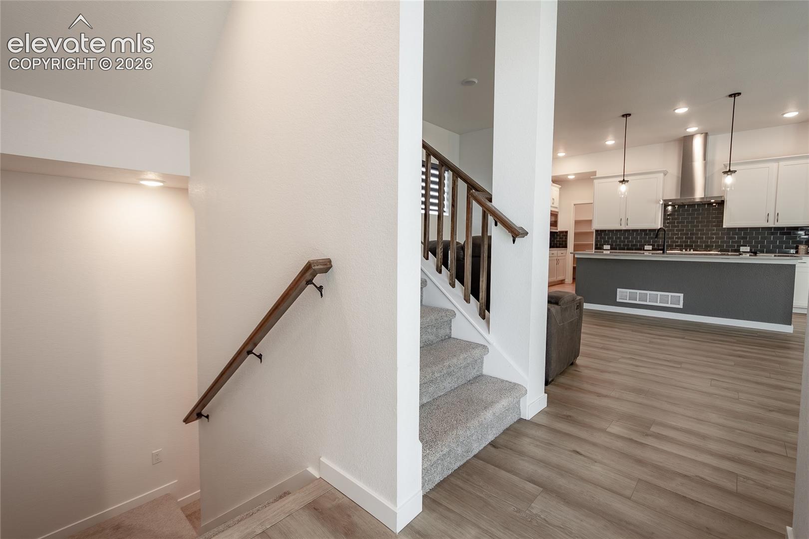 Staircase featuring wood-finish handrails and carpeted treads, wood-finish flooring, kitchen island, white cabinetry, and pendant lighting
