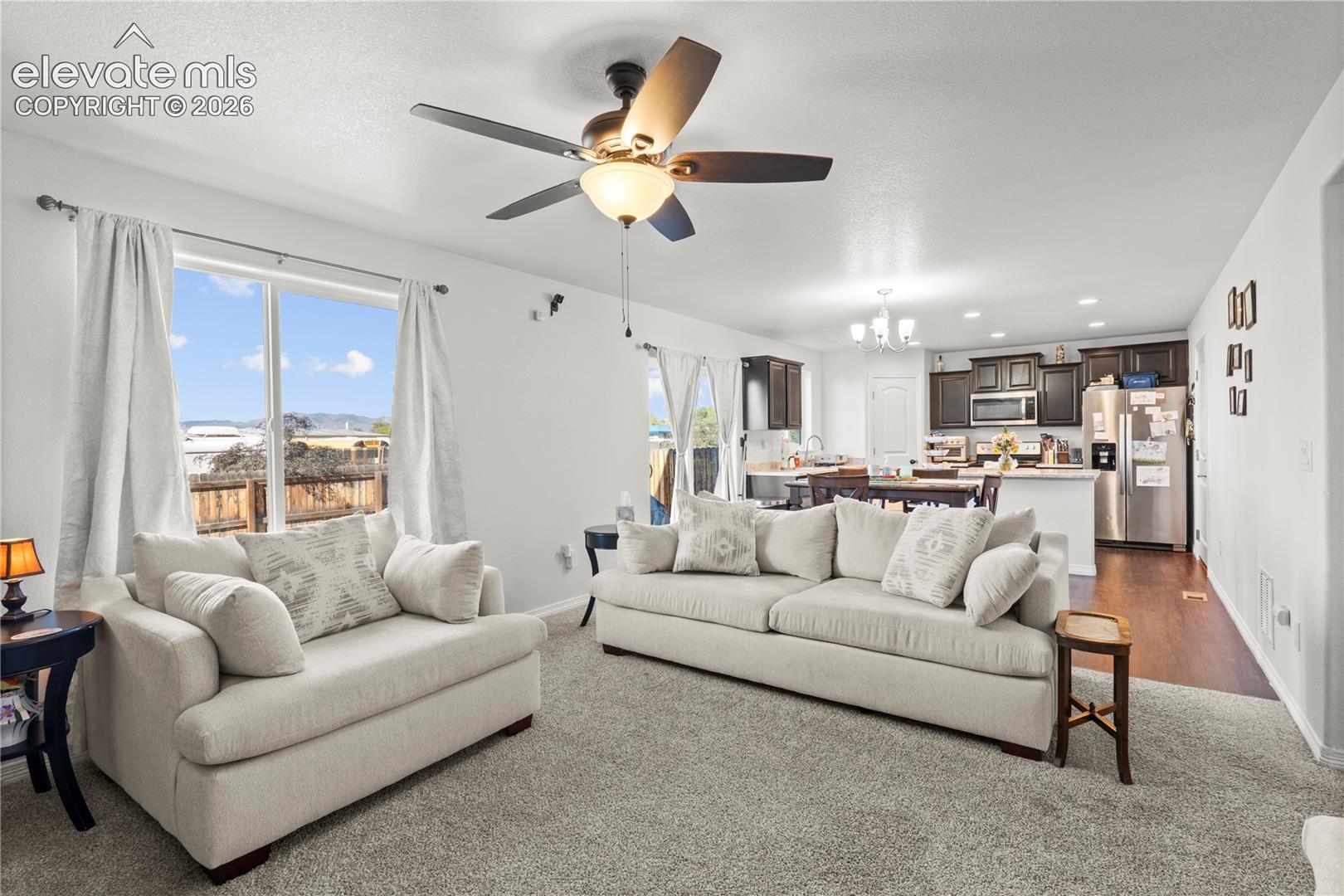 Open-concept interior featuring wall-to-wall carpeting, a ceiling fan with light fixture, large picture windows, and kitchen cabinetry with a peninsula